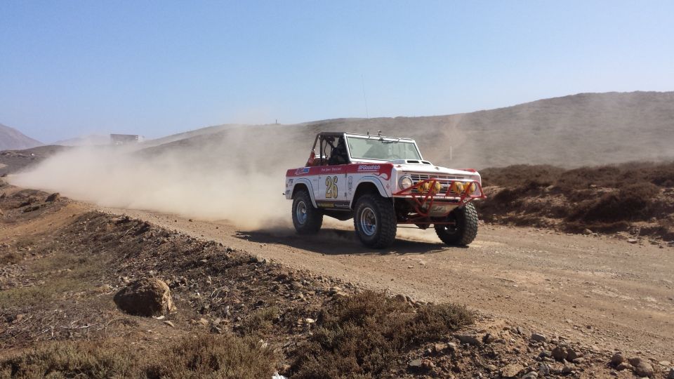 White Ford Bronco with red roll cage and racing decals kicking up dust on a dirt road