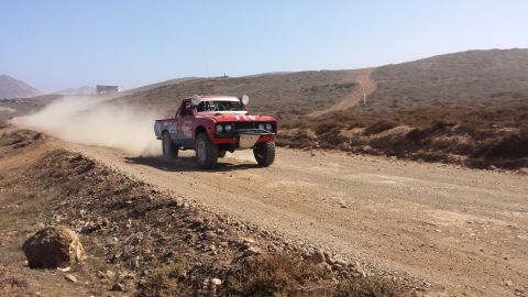 1996 Ford Bronco - A modified red truck races down a dusty desert road under a clear blue sky.