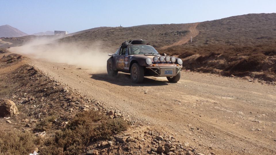Brown rally car with multiple lights on front, kicking up dust on a dirt road