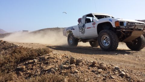 1996 Ford Bronco - A white off-road truck races down a dusty track under a clear blue sky.