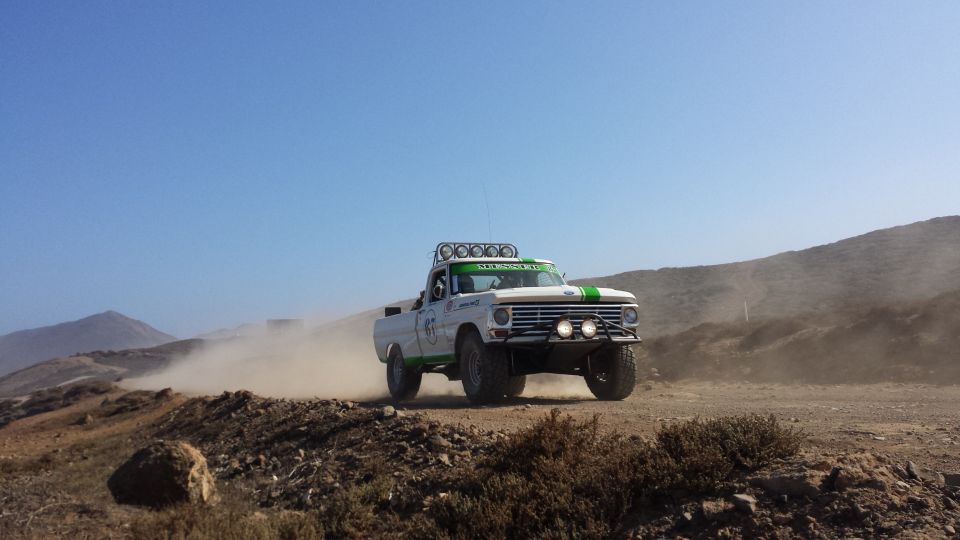 White Ford F-Series truck with green stripes, modified for off-road racing, kicking up dust on a dirt track
