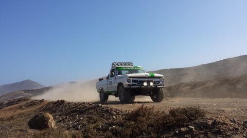 1996 Ford Bronco - A modified Ford F-Series truck races off-road, kicking up a cloud of dust under a clear blue sky.
