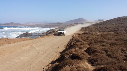 1996 Ford Bronco - A white 1996 Ford Bronco kicks up dust while driving along a coastal road near the ocean.