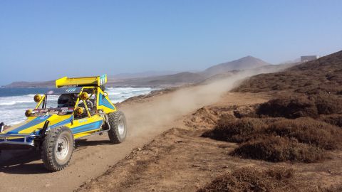 1996 Ford Bronco - An off-road buggy races along a coastal dirt path, leaving a trail of dust in its wake under a clear blue sky.