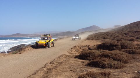 1996 Ford Bronco - Two dune buggies traverse a dusty coastal road under a clear blue sky.