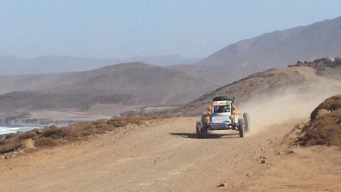 1996 Ford Bronco - An off-road buggy races down a dusty dirt track, with mountains in the background and the ocean visible to the left.
