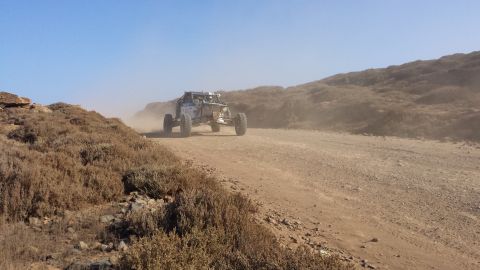 1996 Ford Bronco - An off-road buggy races down a dirt track, kicking up a cloud of dust under a clear blue sky.