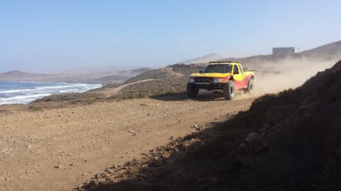 1996 Ford Bronco - A modified truck races along a dusty coastal road, leaving a trail of dust in its wake.