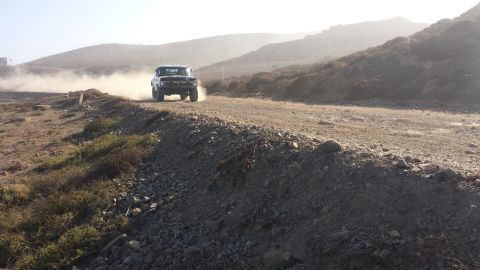 1996 Ford Bronco - A white Ford Bronco kicks up dust as it drives along a dirt road in a hilly, arid landscape.