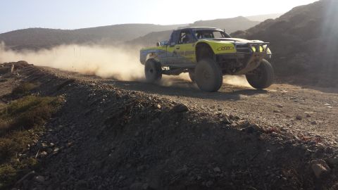 1996 Ford Bronco - A yellow off-road truck races down a dusty dirt track, its large tires kicking up a cloud of dust behind it.