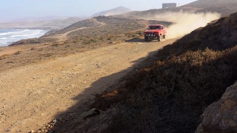 1996 Ford Bronco - A red pickup truck races down a dusty coastal road, leaving a trail of dust in its wake.