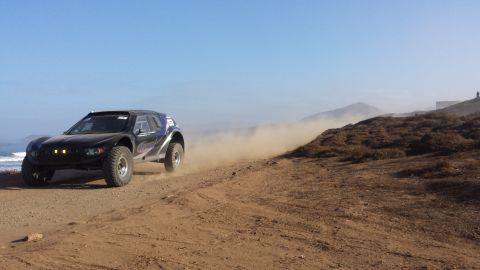 1996 Ford Bronco - A black off-road race truck speeds along a dirt track, kicking up a cloud of dust under a clear blue sky.