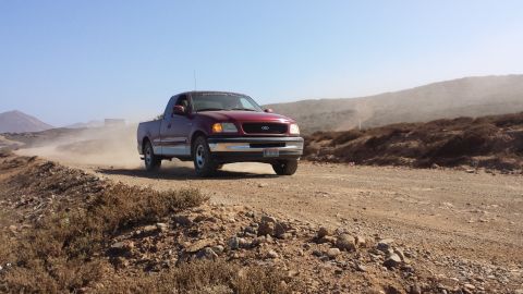 1996 Ford Bronco - A red 1997 Ford F-150 pickup truck kicks up dust as it drives along a remote dirt road.