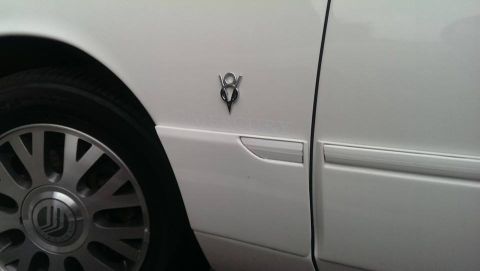 2000 Mercury Grand Marquis - Close-up of a white car's side, showing a V8 emblem and wheel detail.