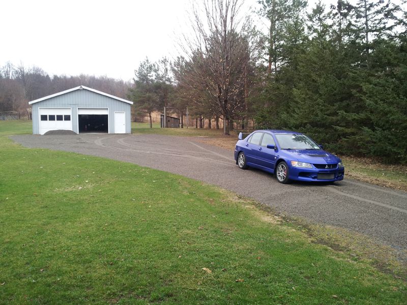 Blue sedan with rear wing parked on gravel driveway near white garage