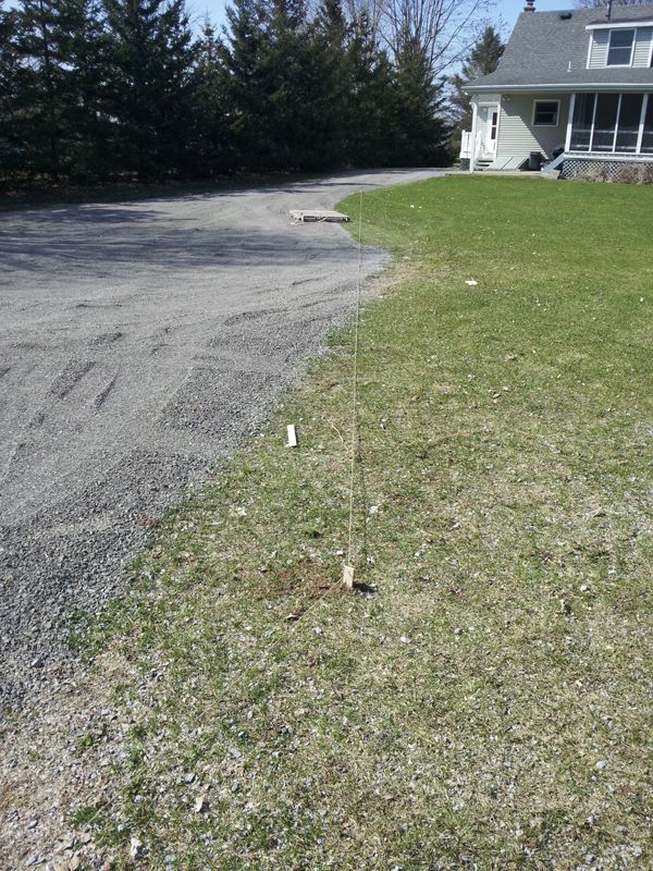 String line staked in grass beside gravel driveway with gray house in background; no vehicle visible.