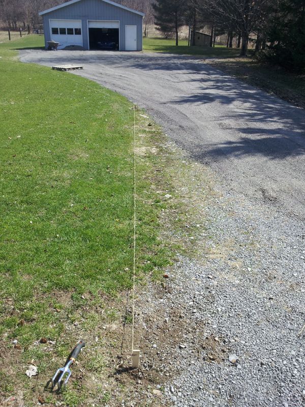 Gravel driveway with string line staked along grass edge, garden fork in foreground, gray garage in background