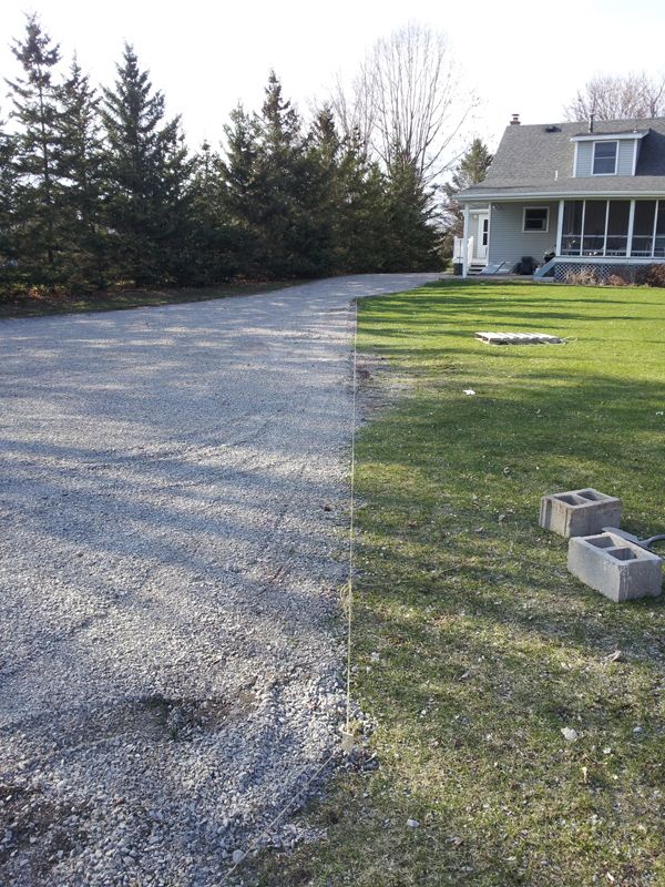 Gravel driveway edged with string line beside a lawn, concrete blocks on grass, blue house and pine trees behind