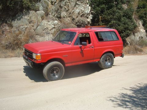 1988 Ford Bronco - This red 1988 Ford Bronco is parked on a dusty, unpaved road with a rocky incline behind it.