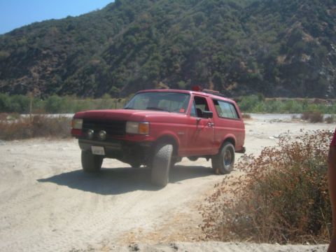 1988 Ford Bronco - A red SUV with off-road tires is parked on a dirt road with a mountainous backdrop.