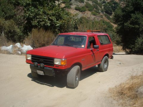 1988 Ford Bronco - This red SUV is parked on a dirt road with hills and trees in the background. It has off-road tires and a roof rack.