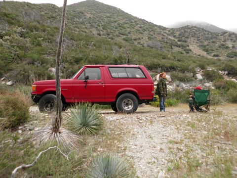 1988 Ford Bronco - A red 1988 Ford Bronco is parked on a dirt road, with a person standing nearby and mountains in the background.