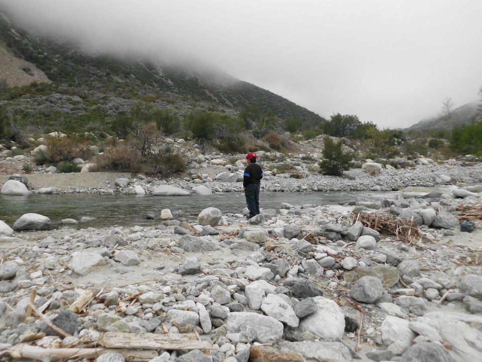 Person in red hat stands in a rocky riverbed with mountains in the background under a foggy sky