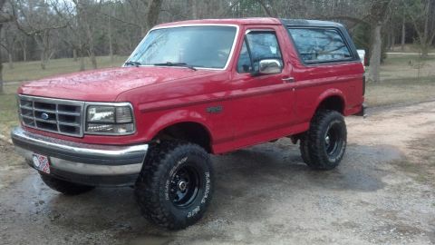 1992 Ford Bronco - This red Ford Bronco is shown from a three-quarter front angle, parked on a dirt road.
