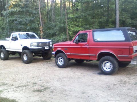 1992 Ford Bronco - Two lifted Ford trucks, a red Bronco and a white F-150, are parked side-by-side on a gravel lot with trees in the background.