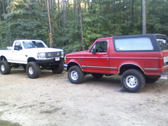 Red 1992 Ford Bronco with black top, lifted on large tires, parked next to a white lifted Ford F-150 truck
