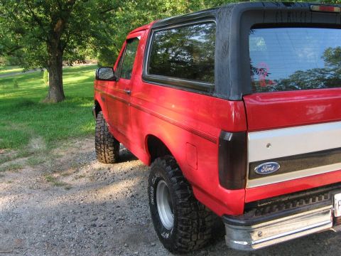 1992 Ford Bronco - Rear quarter view of a red Ford Bronco with a black soft top, parked on a gravel surface near trees.
