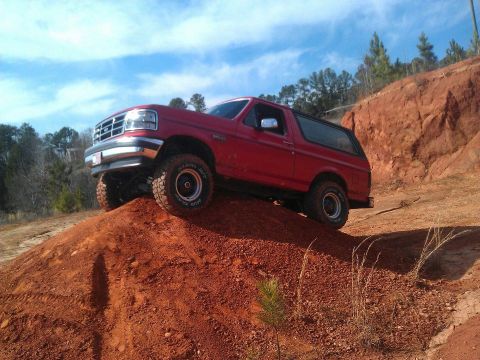 1992 Ford Bronco - A red 1992 Ford Bronco navigates an off-road trail, climbing a steep dirt incline under a partly cloudy sky.