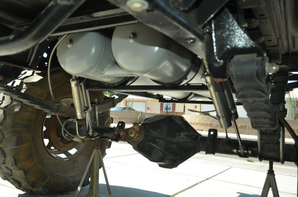 Underside of a vehicle showing leaf springs, shocks, rear axle, and two large propane tanks mounted above the axle