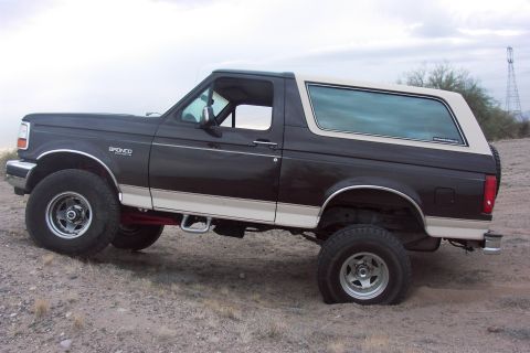 1992 Ford Bronco - Driver-side profile of a dark brown lifted Ford Bronco with white removable hardtop, tan two-tone rocker stripe and Bronco fender badge, sh…