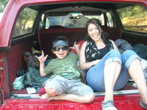 1988 Ford Bronco - Two people are sitting in the cargo area of a red SUV, with camping supplies around them.