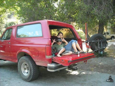 1988 Ford Bronco - Two people relax in the open tailgate of a red Ford Bronco, with a spare tire mounted on the back.