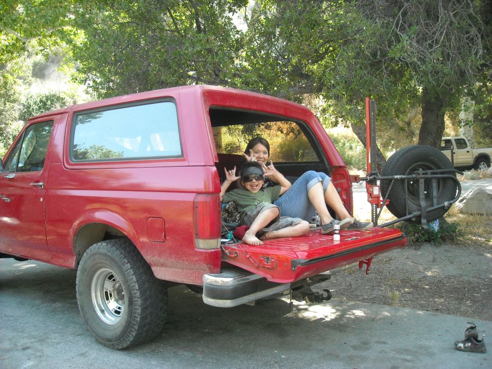 Red Ford Bronco with tailgate open, two people sitting inside. Spare tire mounted on rear. Another truck visible in background