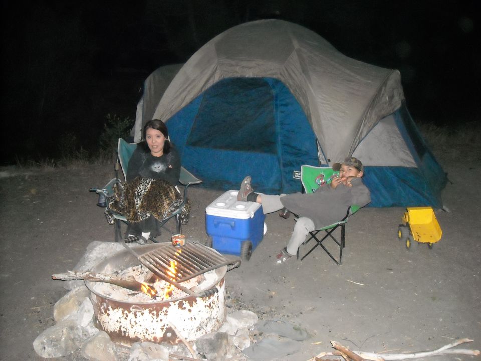 Two people sit in camping chairs by a fire pit and tent at night. A cooler and toy dump truck are nearby