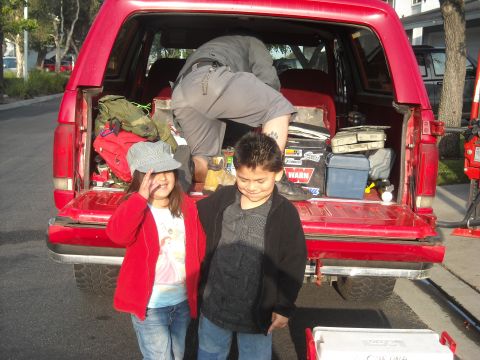 1988 Ford Bronco - A red 1988 Ford Bronco's tailgate is open, revealing camping gear, as two children pose for a photo.