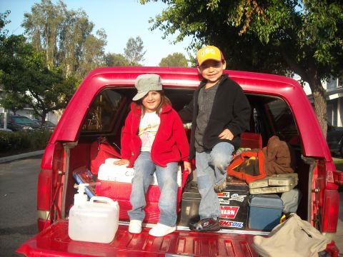 1988 Ford Bronco - Kids are ready for a camping trip, sitting in the back of a red pickup truck filled with supplies.