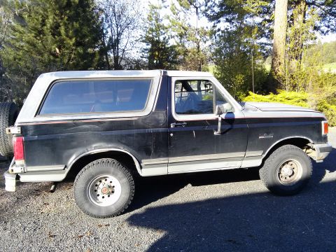 1990 Ford Bronco - Side view of a black 1990 Ford Bronco with a silver top, parked on a gravel lot.