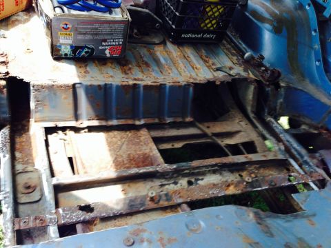 1979 Ford Bronco - The interior floor pan of a vehicle shows extensive rust damage, with a box of red jacks and a milk crate visible.