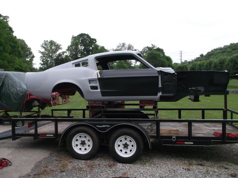 1967 Ford Mustang - A car body, likely a 1967 Mustang fastback, is being transported on a trailer, showing its unfinished state.