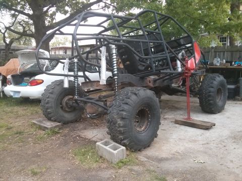 1986 Ford Bronco - A custom-built off-road buggy frame is shown with large tires and a red jack supporting the rear, with a white sedan in the background.
