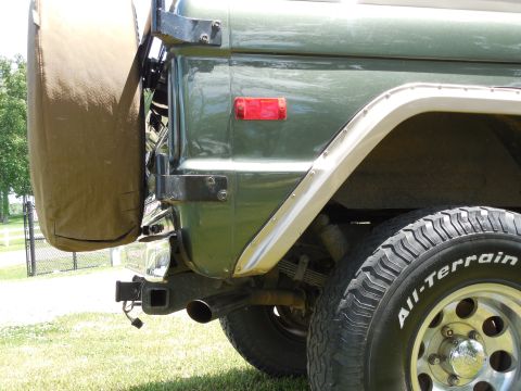 1975 Ford Bronco - Close-up of the rear corner of a green vintage Ford Bronco showing the tan-covered swing-out spare tire and its black hinge brackets, red s…