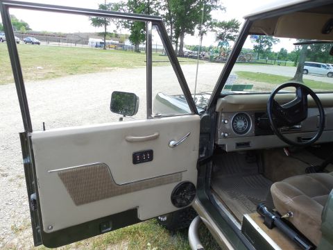 1975 Ford Bronco - Open driver's door of a dark green early Ford Bronco showing a tan door panel with cloth insert, chrome trim, an added rocker-switch panel…