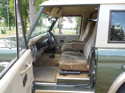 1975 Ford Bronco - Interior view of an early Ford Bronco shot through the open driver's door, showing tan/brown bucket seats, a black steering wheel with roun…