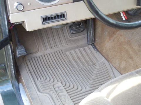 1975 Ford Bronco - Driver's footwell of a vintage Ford Bronco showing a tan Husky ribbed rubber floor mat over tan carpet, rubber-pad brake and gas pedals, a…