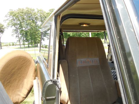 1975 Ford Bronco - Interior view through an open door of a green vintage Bronco showing tan cloth seats, the rear bench upholstered with a woven pine-tree and…