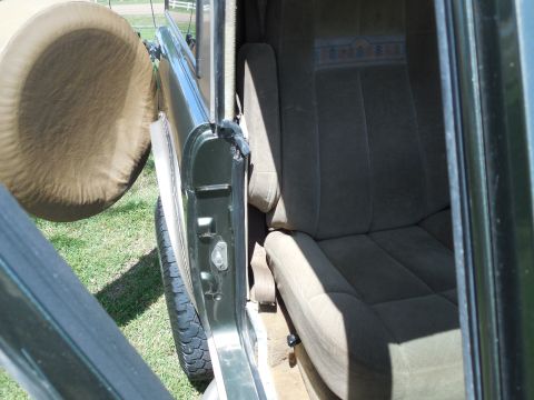 1975 Ford Bronco - Close view into the rear compartment of a green vintage Bronco through the open passenger door, showing the tan cloth rear bench with a wov…